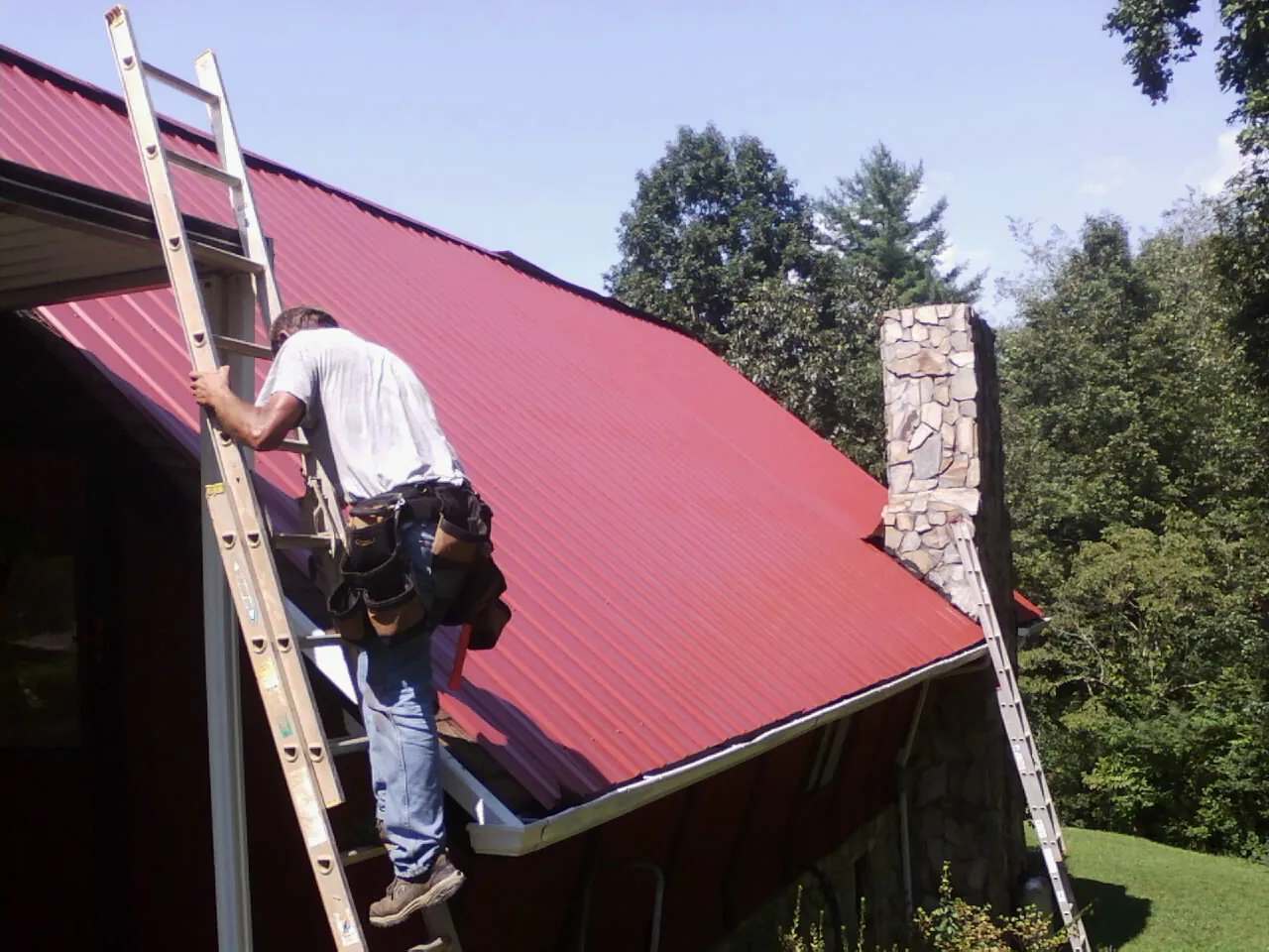 Roofer on a ladder beside a red metal roof and stone chimney — Soffit & Fascia Repair by Roofing Brothers in Saint Paris